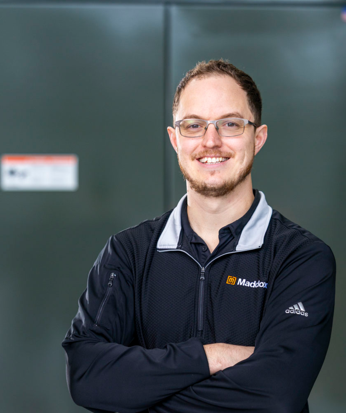Smiling man with glasses wearing a black Maddox Adidas jacket, arms crossed against a gray background.