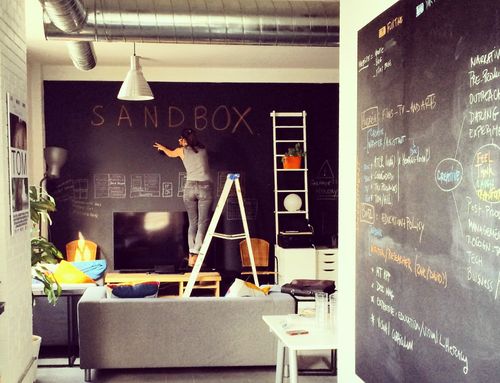 A man and a women smiling on a couch with sketches and laptops in front of them