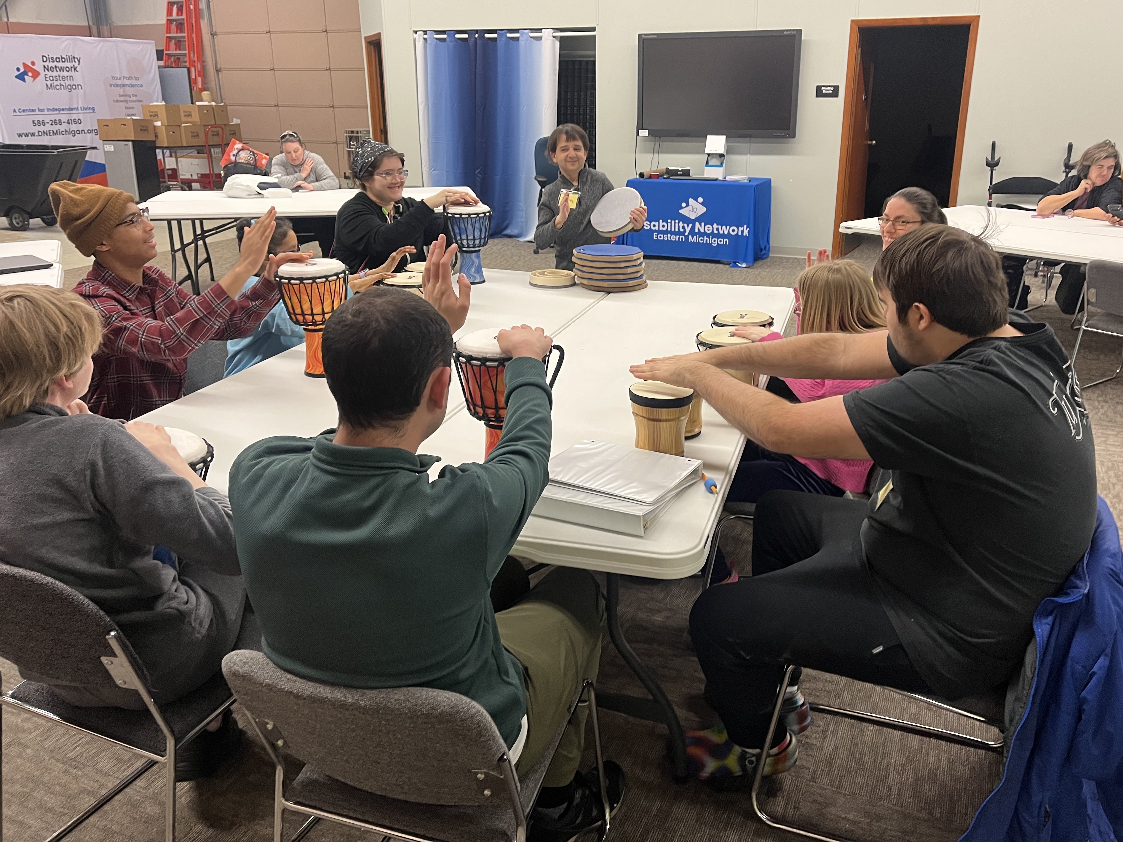 Nine people are gathered around a table playing percussion instruments at Disability Network Eastern Michigan's Troy office.