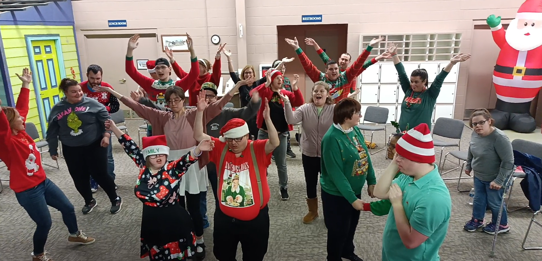 A diverse group of people dressed in holiday outfits is dancing together at Disability Network Eastern Michigan's Holiday Karaoke Party.