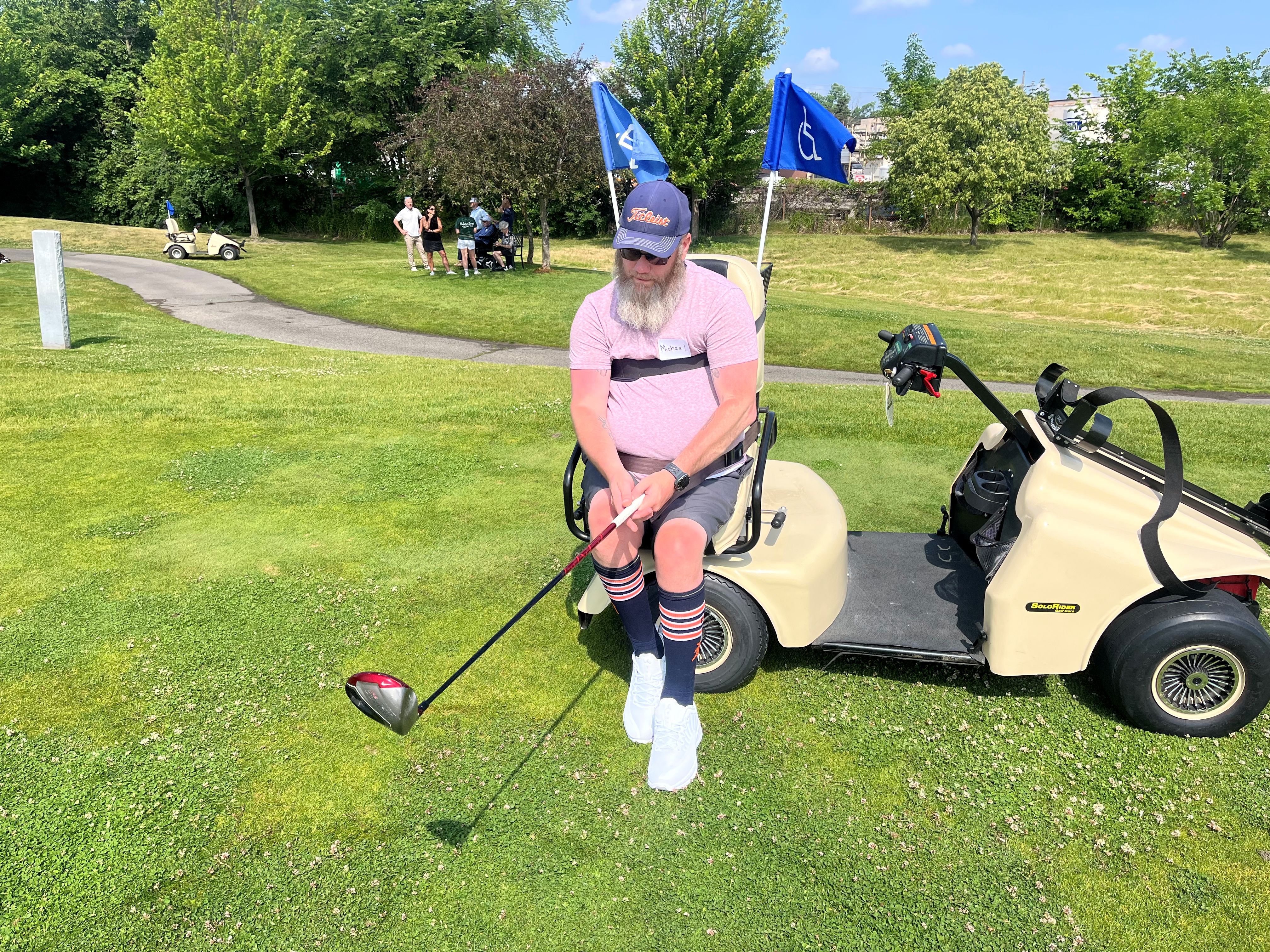 A man is using an adaptive golf cart to help him stand when hitting a golf ball during a Disability Network Eastern Michigan adaptive golf activity.