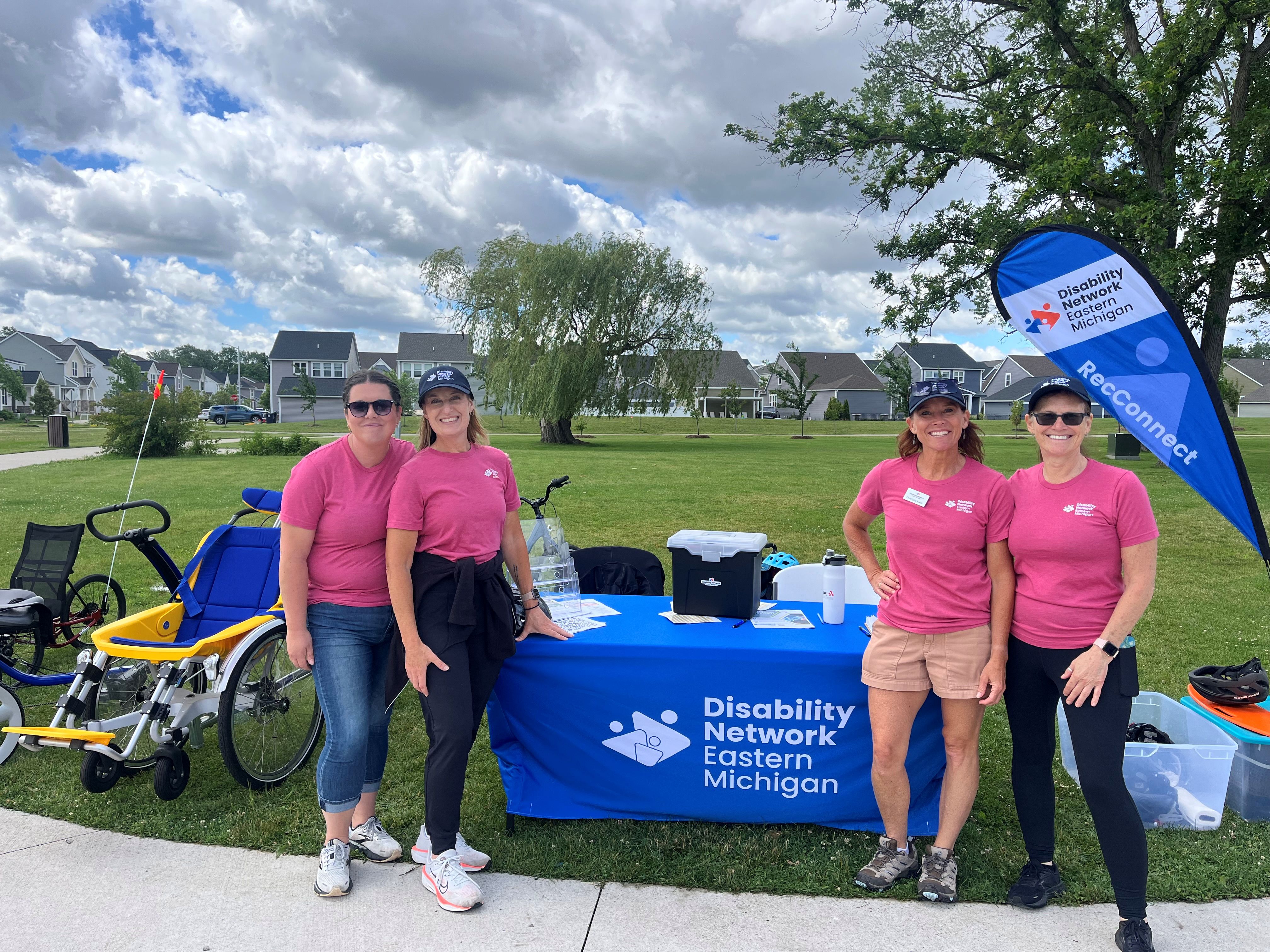 Smiling Disability Network Eastern Michigan staff stand in front of a registration table next to adaptive bikes, ready to greet adaptive recreation participants.