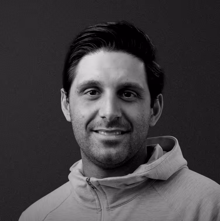Portrait of a smiling man with dark hair wearing a light-colored zip-up hoodie against a dark background.