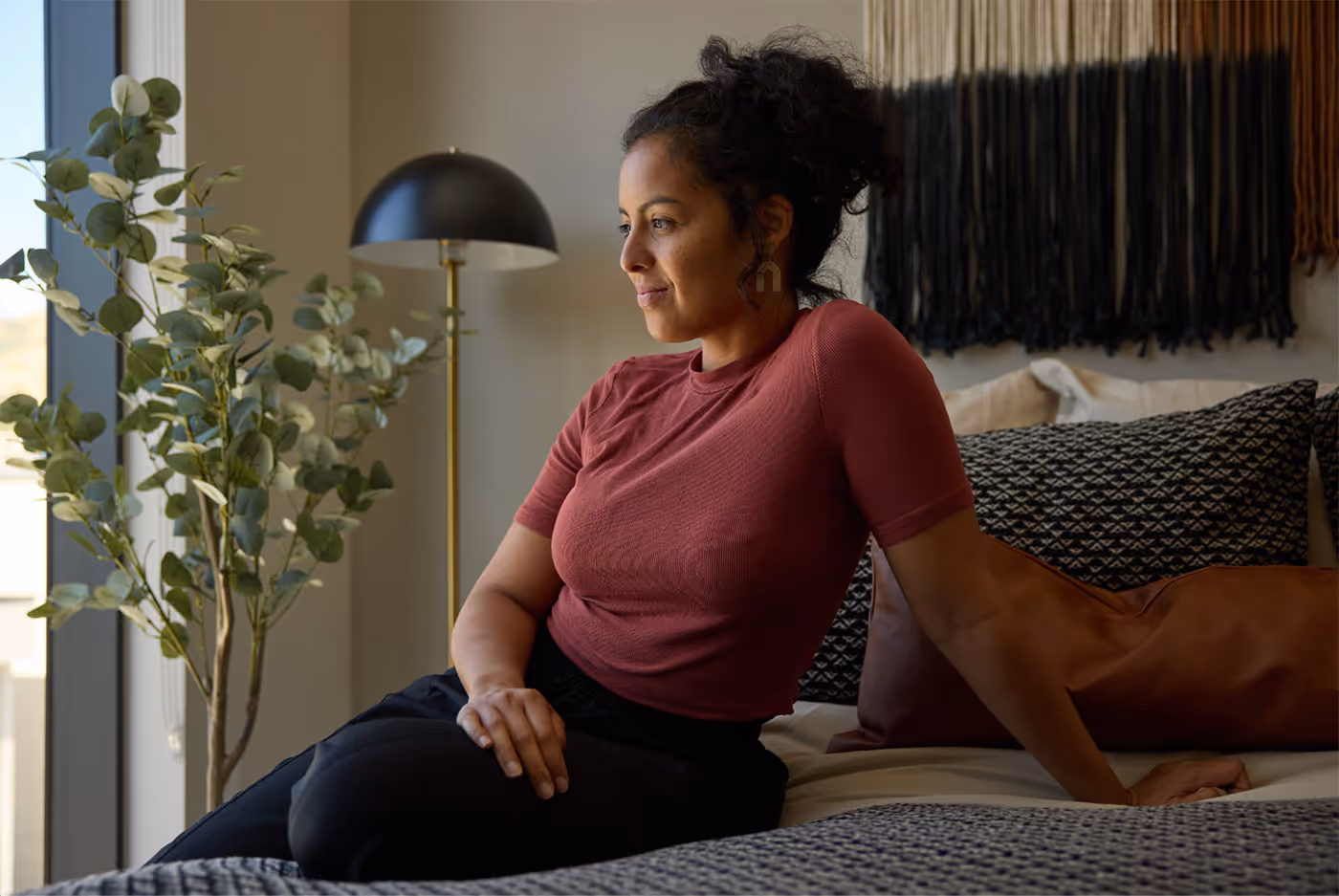 Woman with curly hair in a red shirt sitting on a bed next to patterned pillows and a leafy plant by a window.