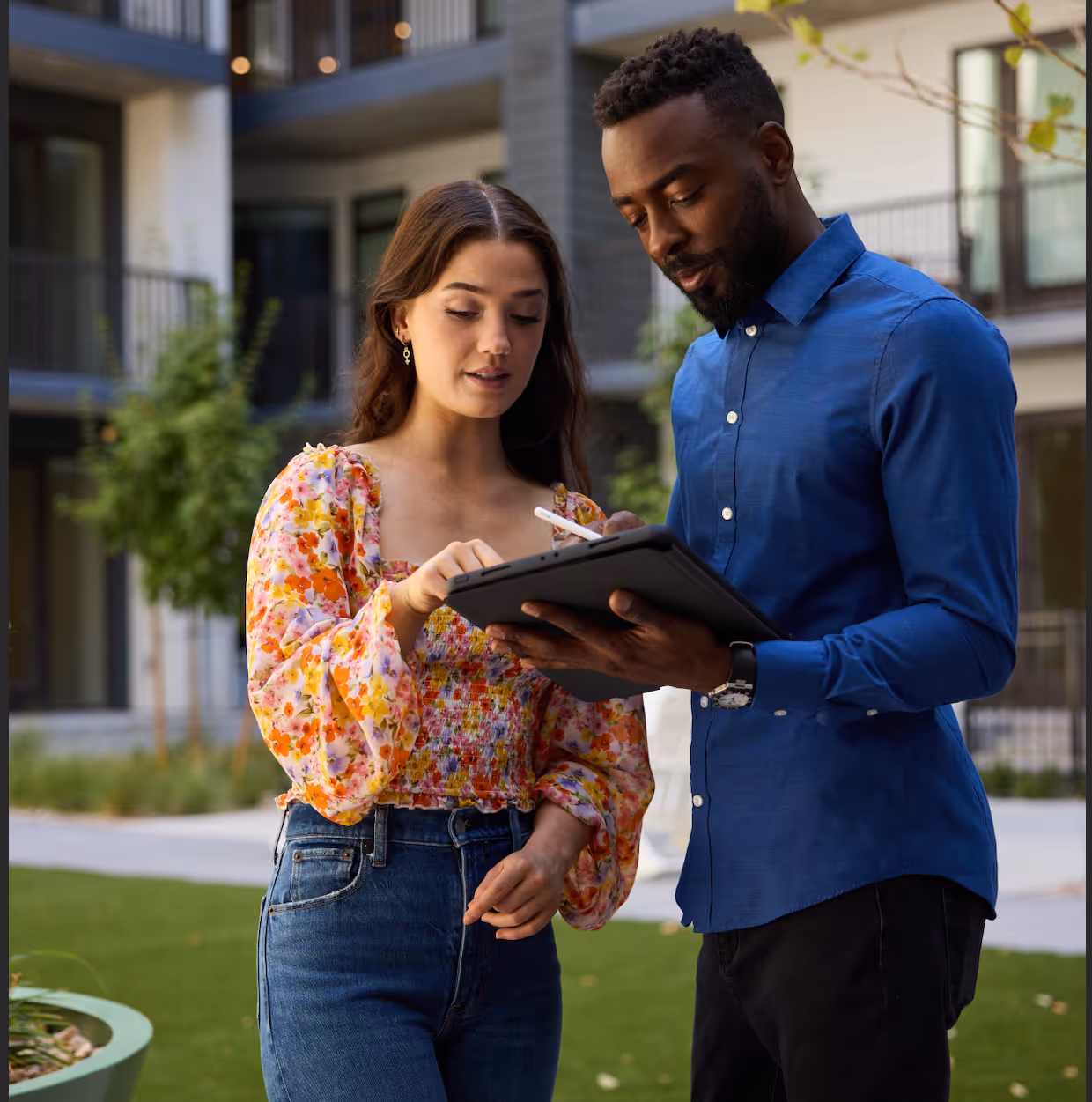 A woman in a floral blouse and jeans is using a stylus on a tablet held by a man in a blue shirt, outdoors in a residential area.