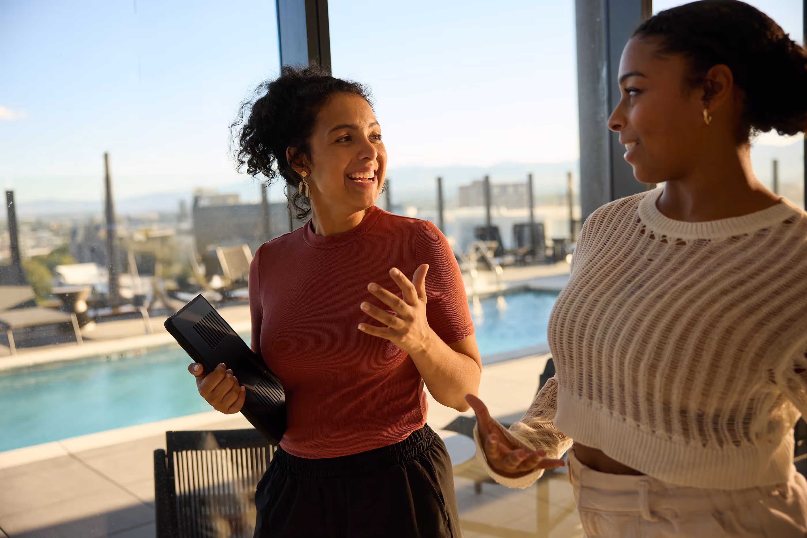 Two women smiling and talking indoors near a large window overlooking a pool.