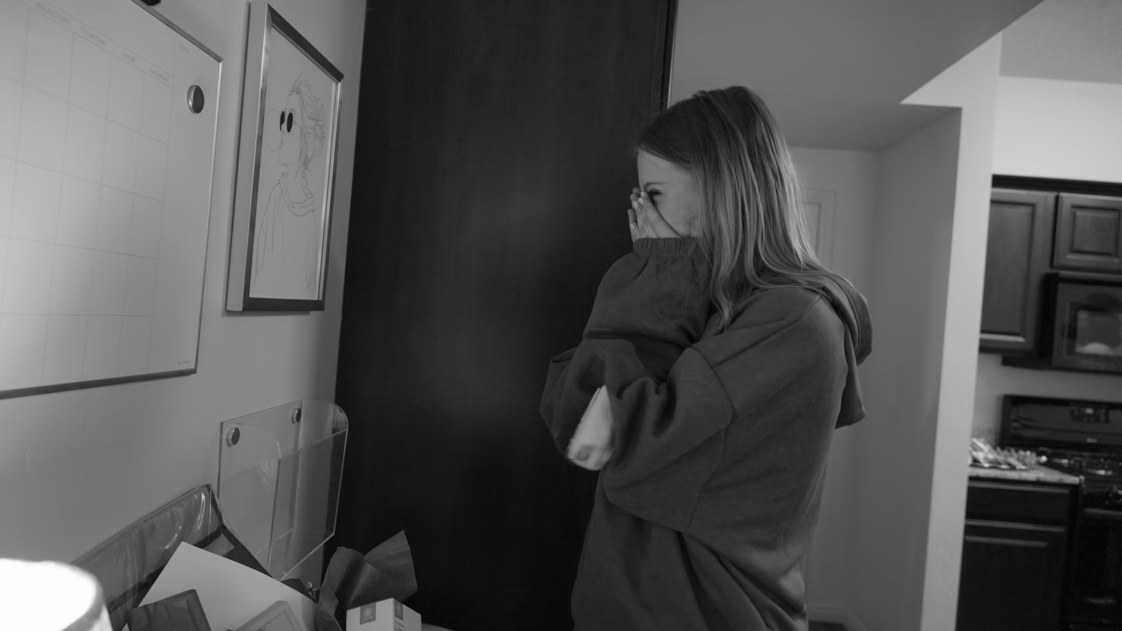 Young woman in hoodie covering her face with hands, standing in a home kitchen area.