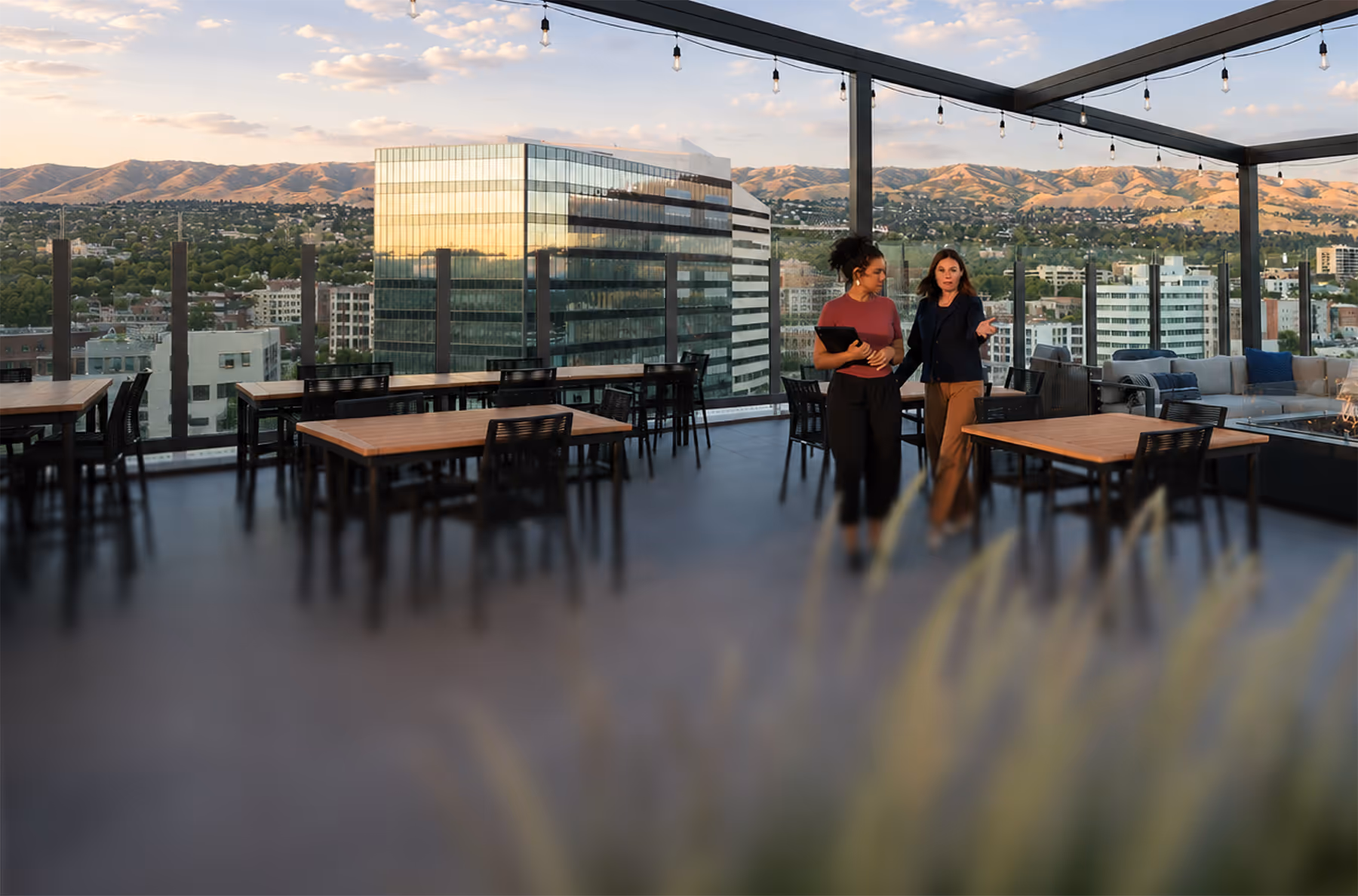 Two women talking on a rooftop patio with wooden tables and cityscape and hills in the background.