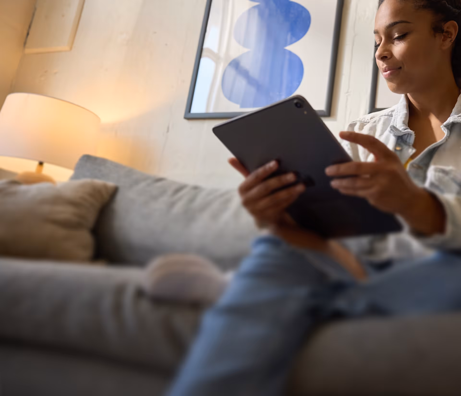 Young woman in denim jacket sitting on a gray couch using a tablet device.