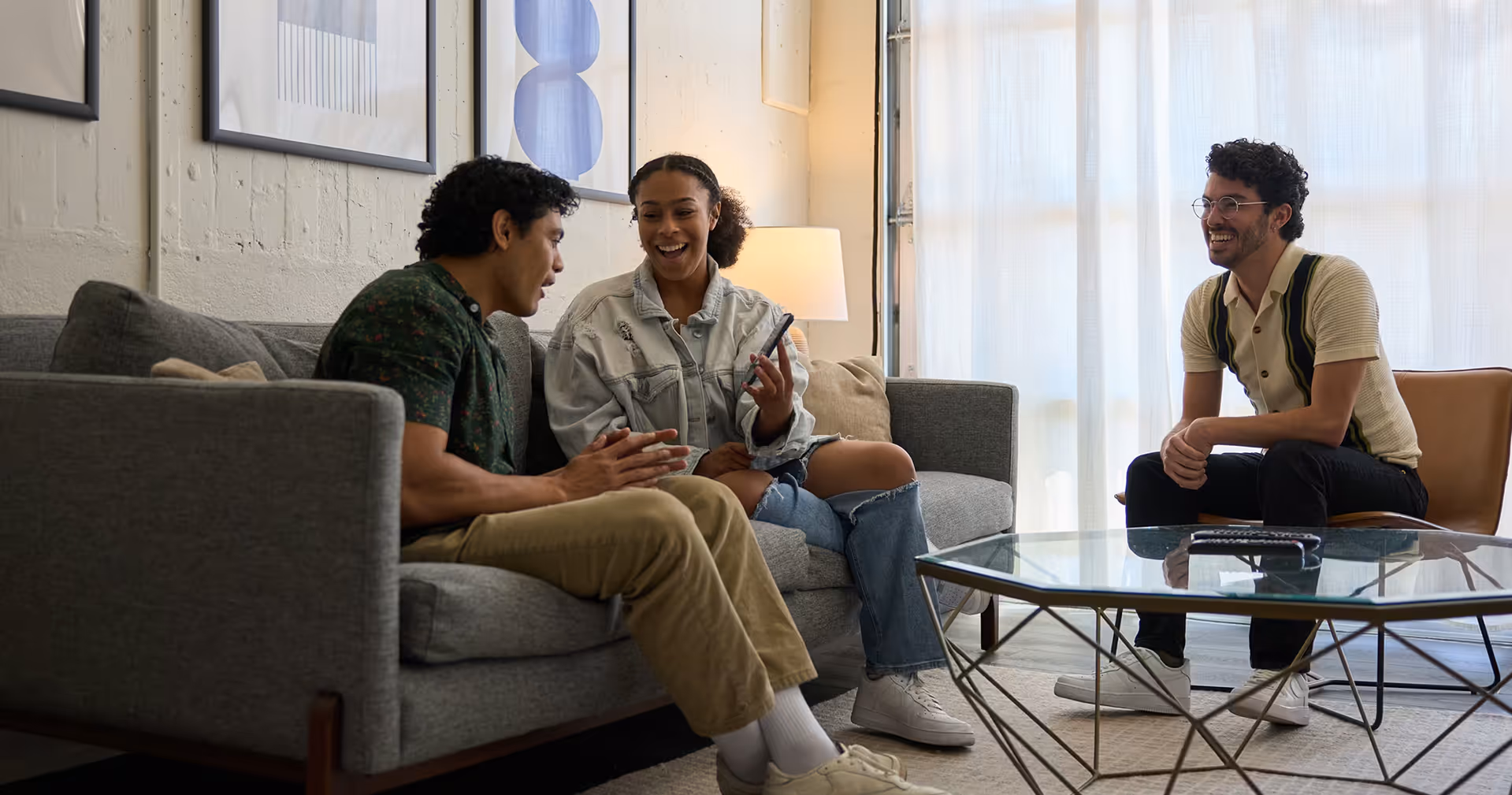 Three people smiling and talking in a living room with a gray sofa and glass coffee table.