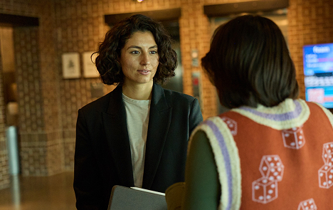 Two women conversing indoors, one holding a folder, with a brick wall in the background.