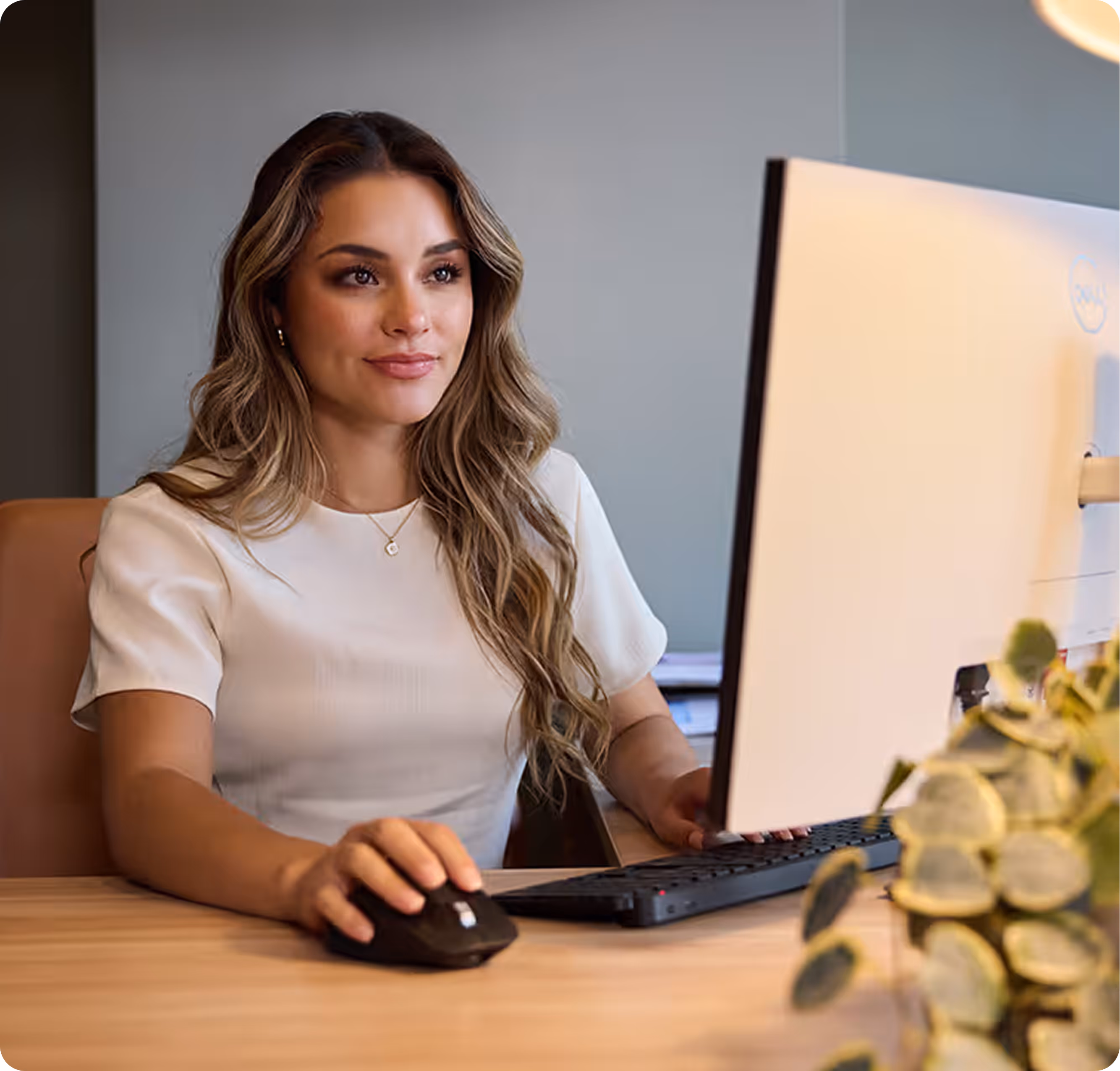 Woman with long hair sitting at desk using a computer mouse and keyboard, looking at the monitor.