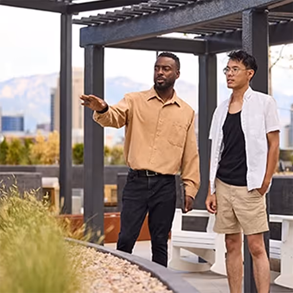 Two men standing outdoors by plants; one gestures while the other listens attentively.