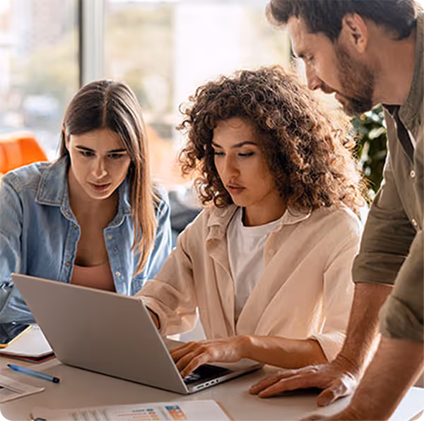Three young adults working together on a laptop at a table in a bright office space.