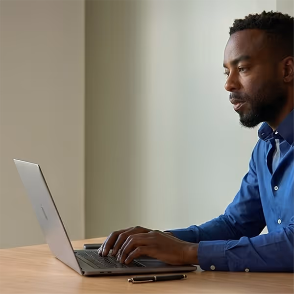 Man wearing blue shirt typing on a laptop at a wooden desk with a pen and phone nearby.