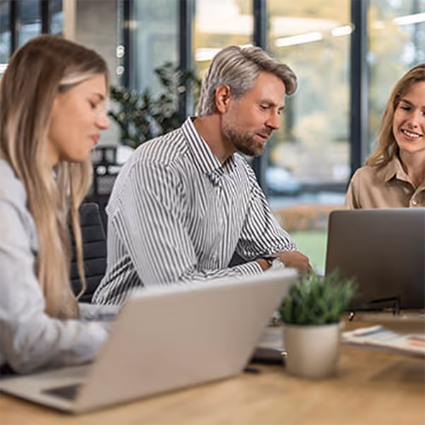 Three professionals working on laptops and discussing at a wooden conference table in an office.