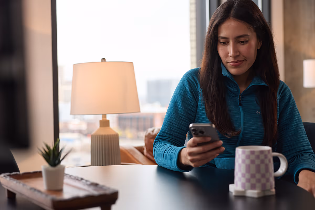 Woman in blue jacket looking at her smartphone sitting at a table with a checkered mug and plant nearby.