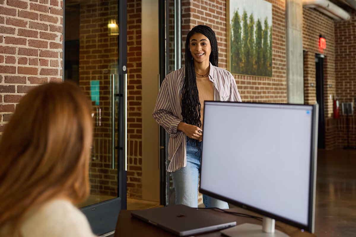 Woman with long braided hair smiling as she approaches another woman at a desk with a computer.