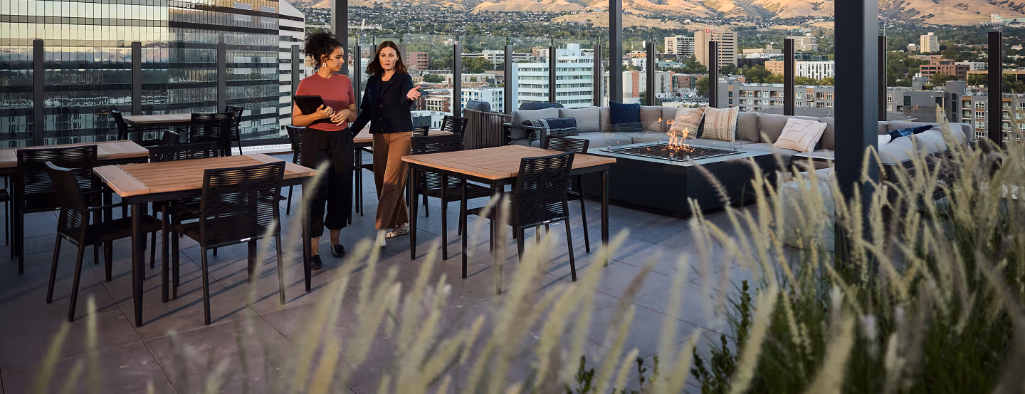 Two businesswomen talk on an outdoor terrace with tables, chairs, and city skyline views.