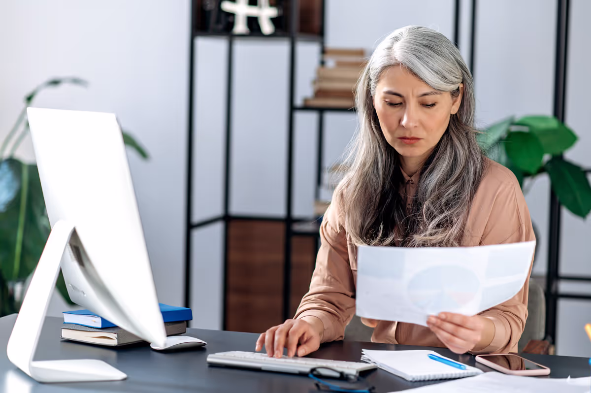 A woman with long gray hair sits at a desk in an office, looking thoughtfully at a printed document while working on a computer.