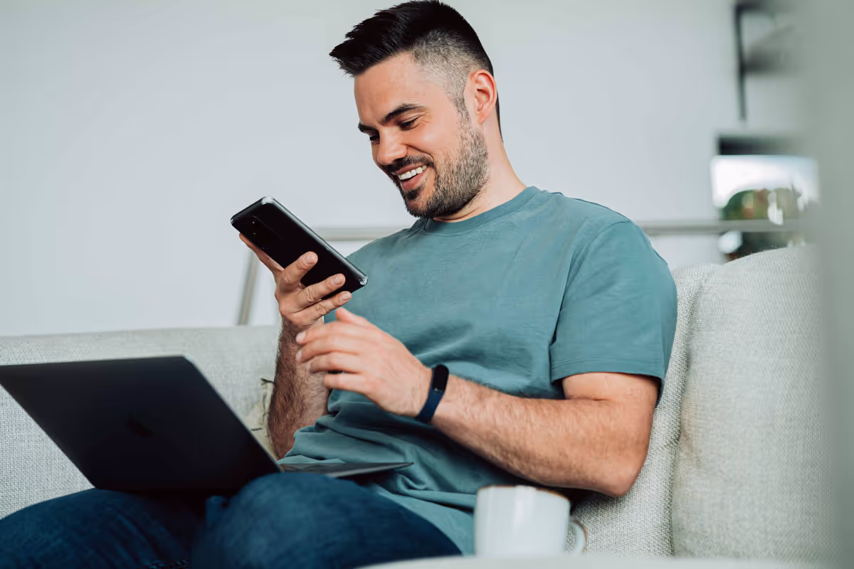 A man sits on a couch using a laptop on his lap and looking at his phone, smiling. A white mug is on the table in front of him.
