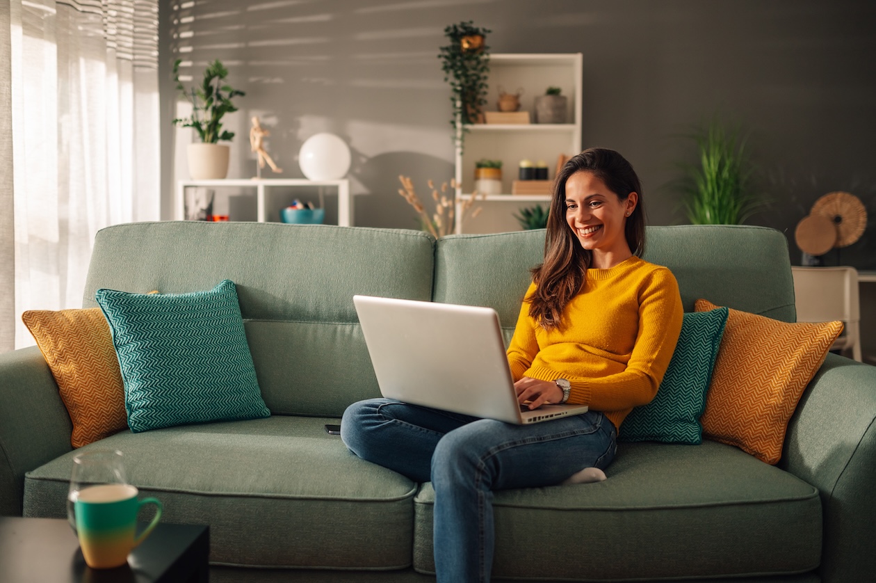 A woman with dark hair sits comfortably on a light green sofa, wearing a yellow sweater and jeans while working happily on a laptop on her lap. Her modern living room has decorative pillows, a coffee table, and bookshelves filled with plants, bathed in natural light filtering through window blinds.