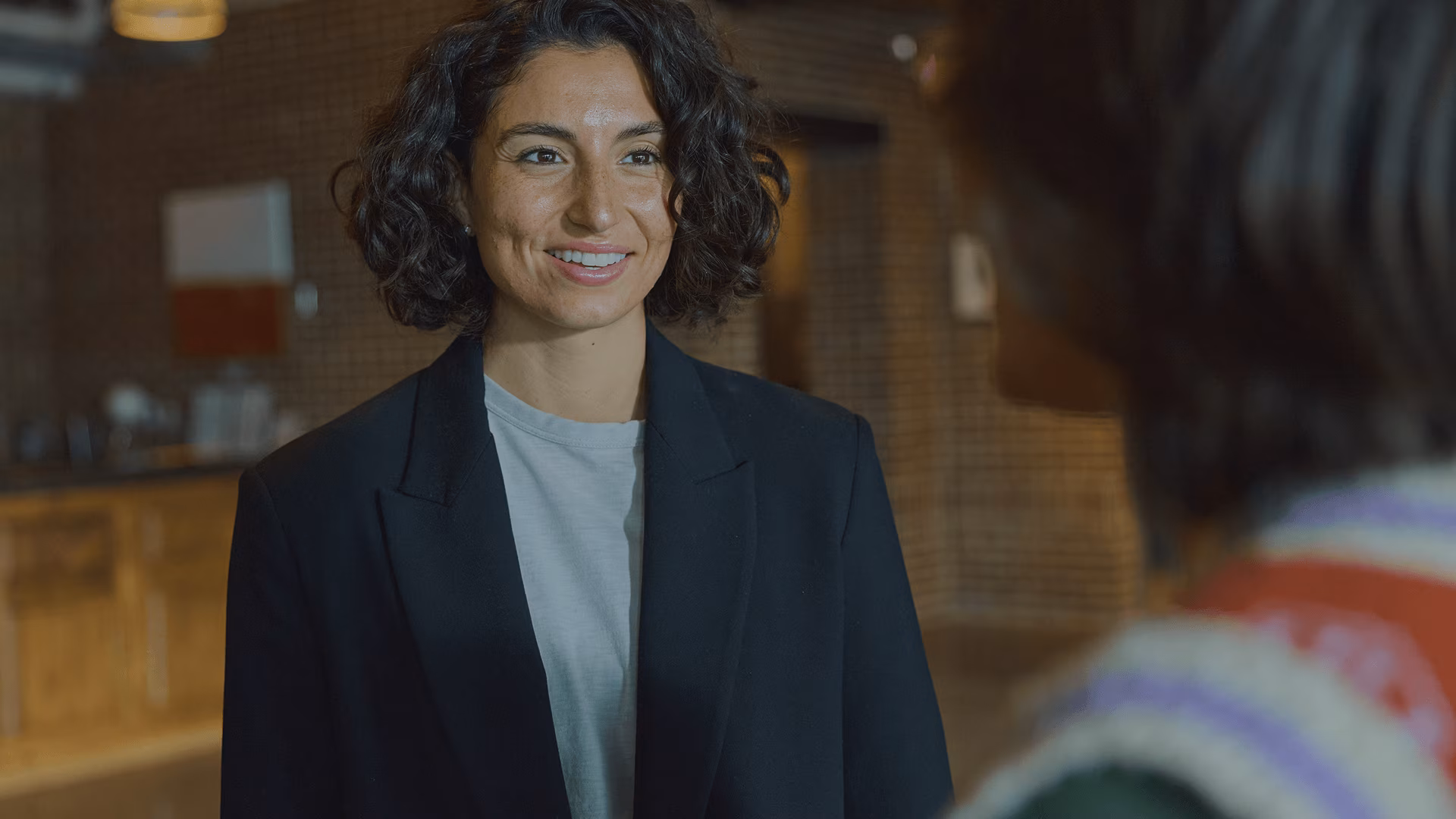 A smiling woman in a black blazer and light-colored shirt speaks with another person in a warmly lit indoor setting