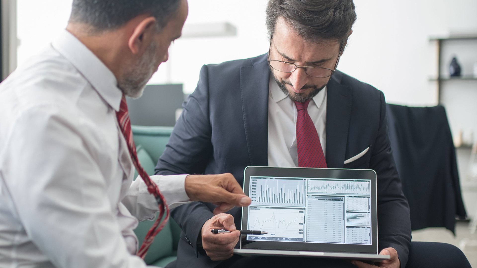 2 people sitting on a couch wearing formal attire looking at a data spreadsheet on a laptop