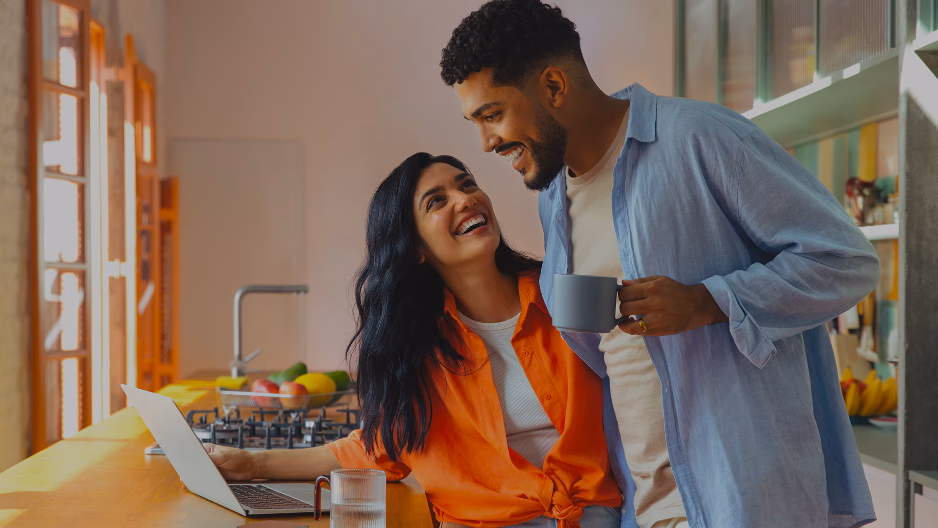 A couple smiles together in their modern apartment kitchen while enjoying coffee next to an open laptop, representing a comfortable and connected rental living experience.