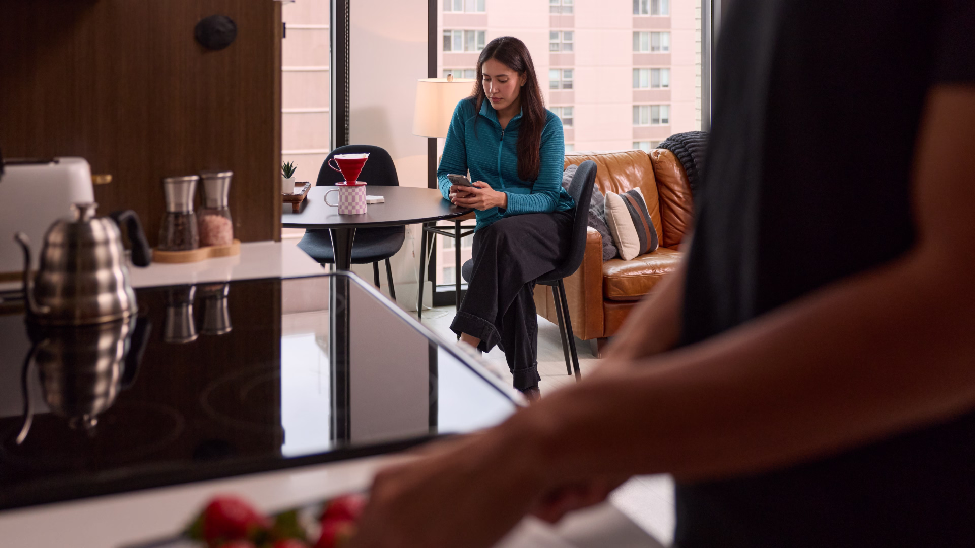 A person utting fruit on a cutting board with another person sitting at a round table in the background with their phone in hand