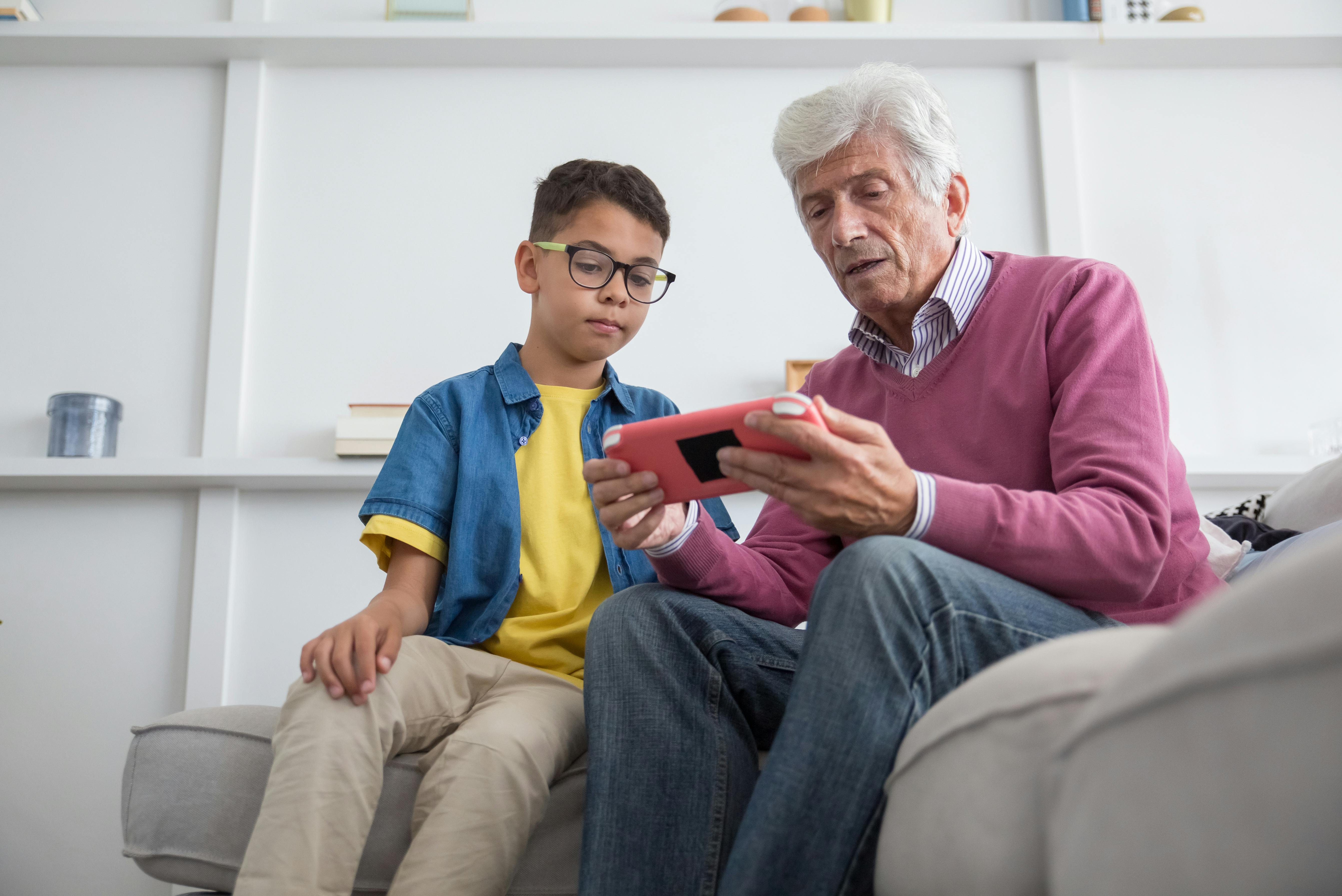 Senior man sitting on a couch showing a smartphone screen to a young boy wearing glasses.