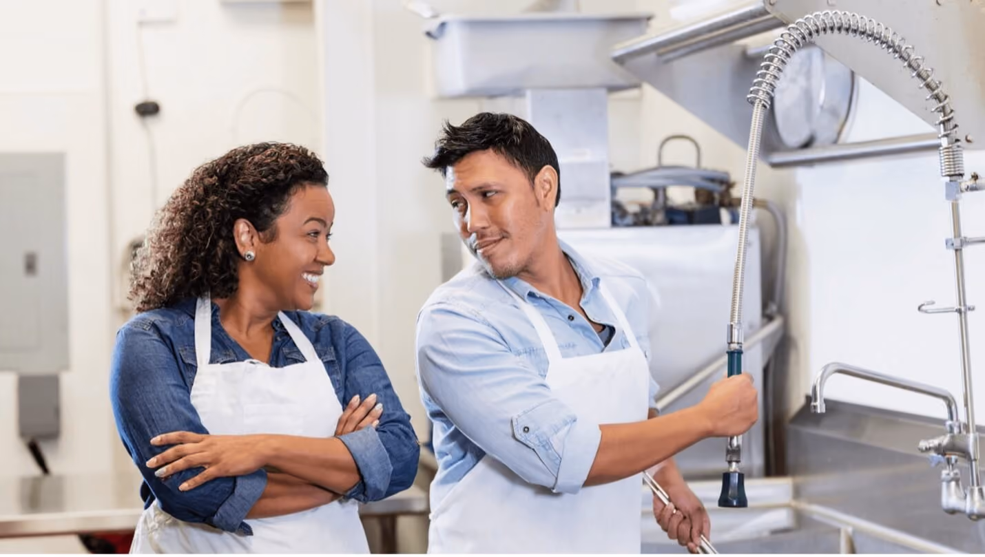 A man and woman wearing aprons stand together in a kitchen, preparing food with a warm and inviting atmosphere.