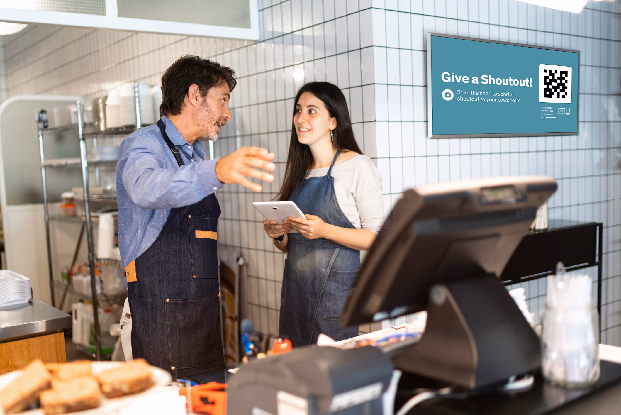 Two individuals wearing aprons stand together in front of a counter, engaged in a conversation