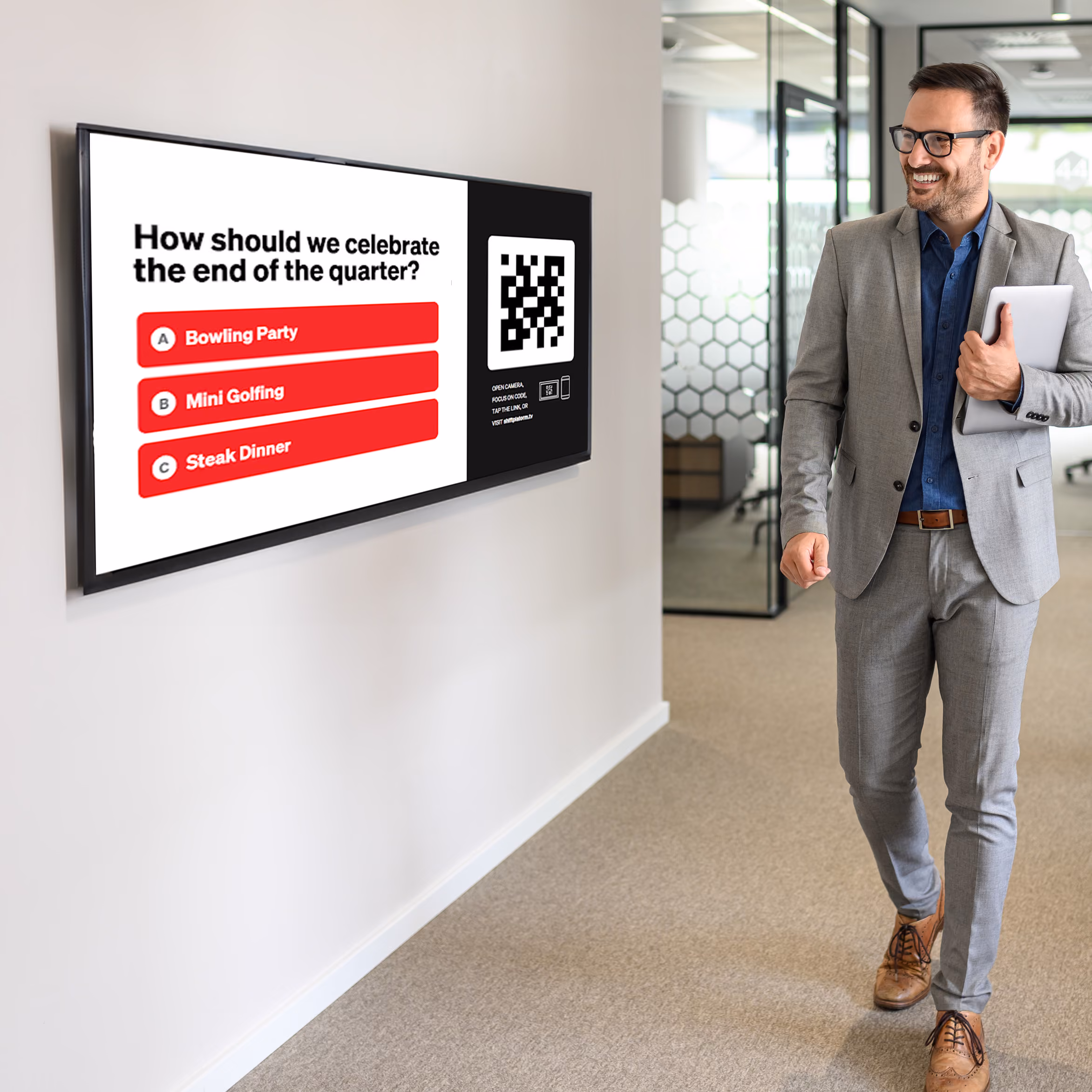 A man walking in an office hallway looking at a digital screen on the wall