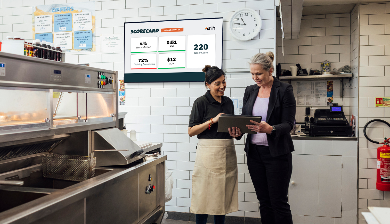 Two women in a kitchen looking at a list with a digital screen in the background