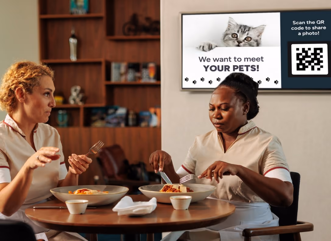 Two women enjoy a meal at a table, with a cat visible on a screen in the background.