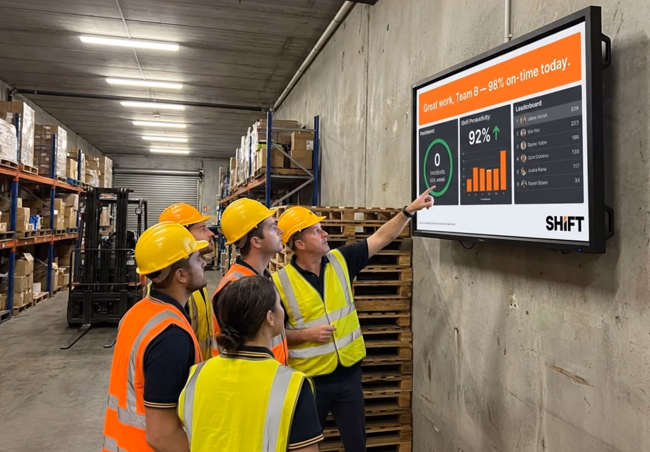 A group of people in a warehouse attentively watching a large screen displaying information or visuals.
