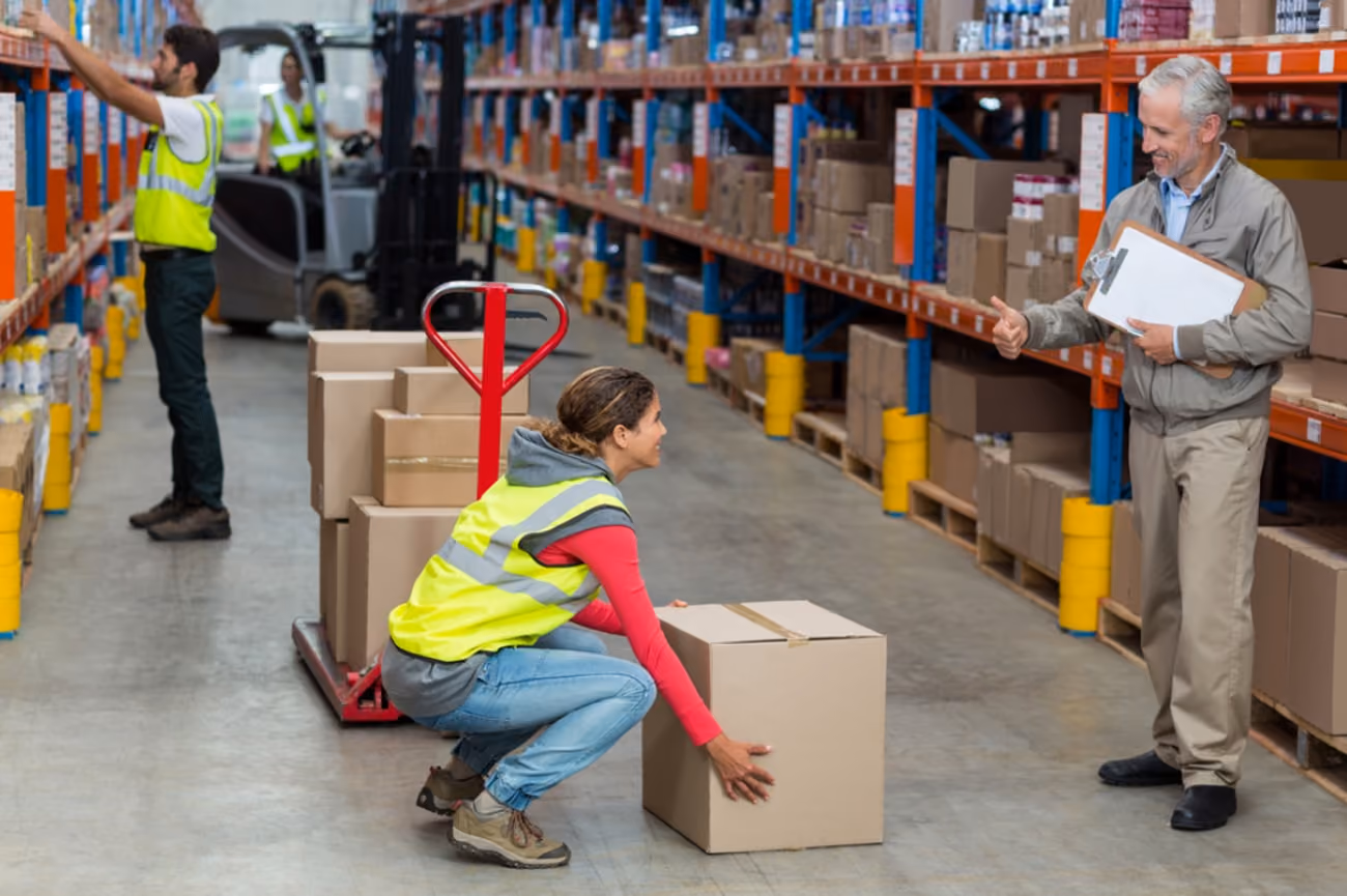 A man and woman stand in a warehouse surrounded by stacked boxes, discussing their work.