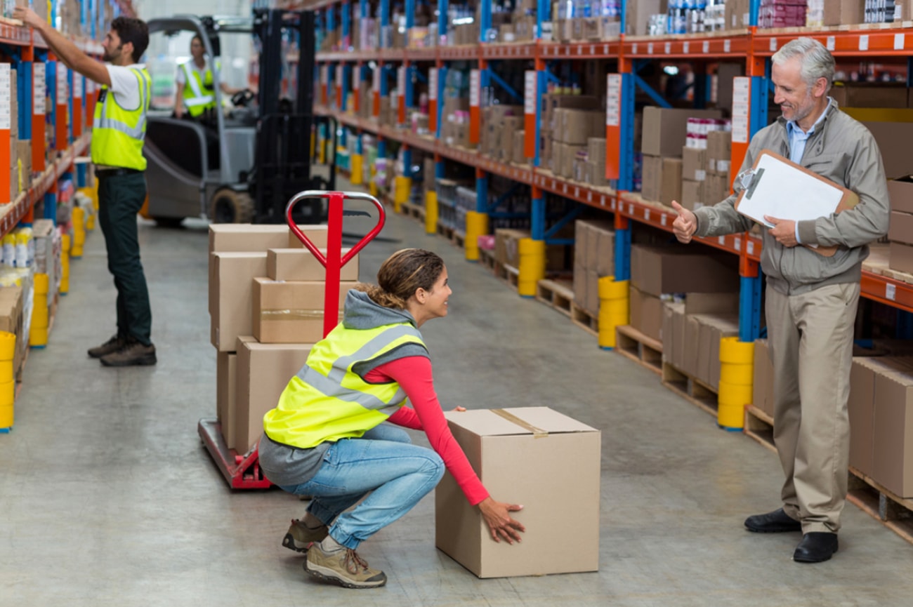 A man and woman stand in a warehouse surrounded by stacked boxes, discussing their work. 