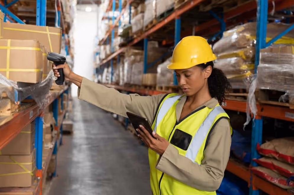 A woman in a hard hat and safety vest scans packages with a tablet, ensuring safety and efficiency in her work environment.