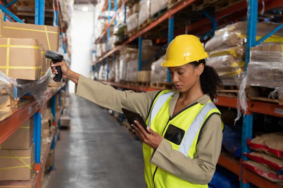 A woman in a hard hat and safety vest scans packages with a tablet, ensuring safety and efficiency in her work environment.  