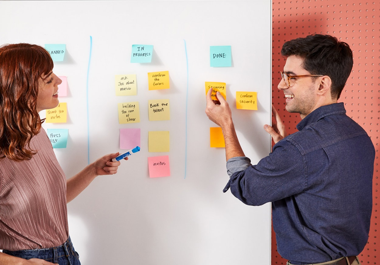 A man and woman stand beside a whiteboard covered in colorful sticky notes, discussing ideas collaboratively.