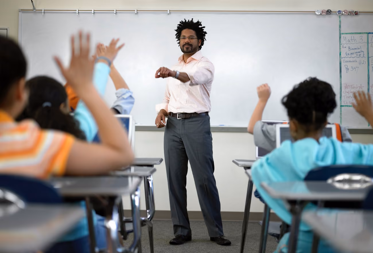 A teacher stands in front of a classroom, raising his hands to engage students in a lesson.