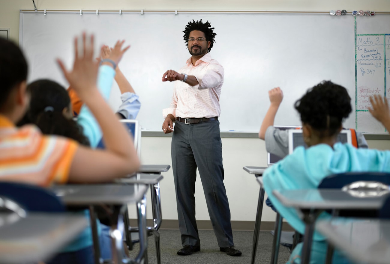 A teacher stands in front of a classroom, raising his hands to engage students in a lesson.