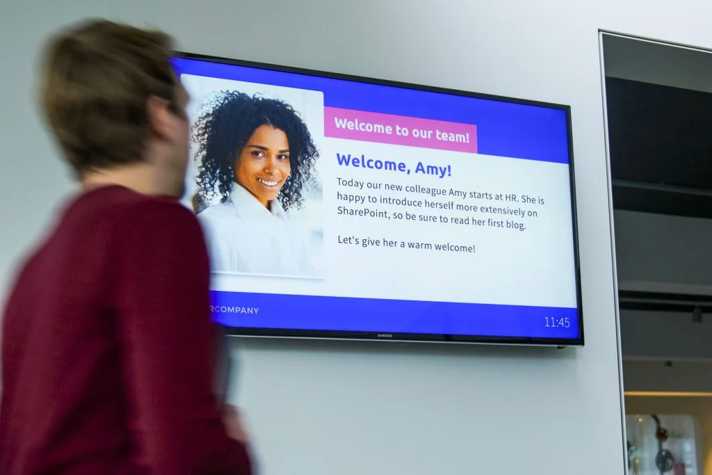 A woman is positioned in front of a TV screen showing a welcome announcement.  
