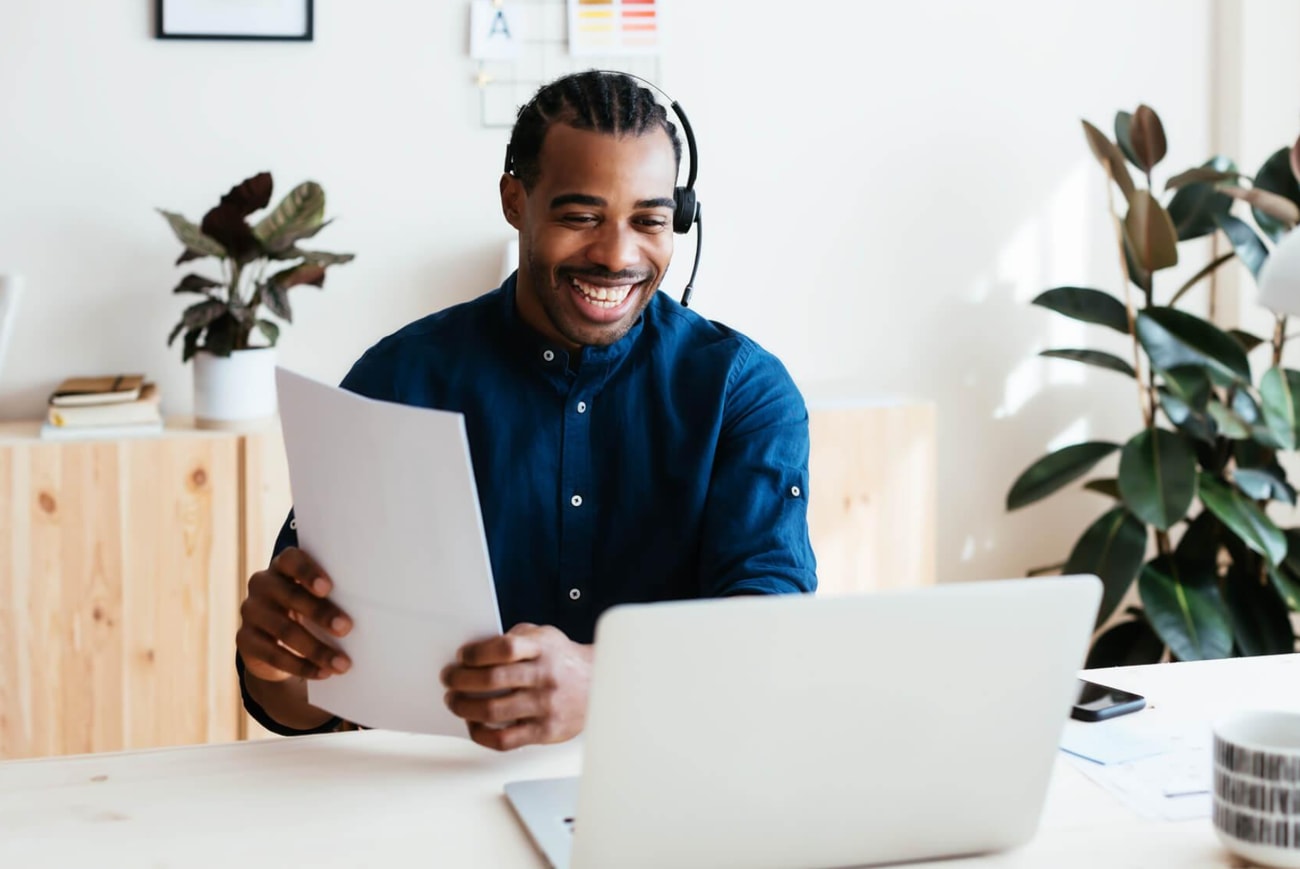 A smiling man sits at a desk with a laptop and papers, appearing engaged and focused on his work.