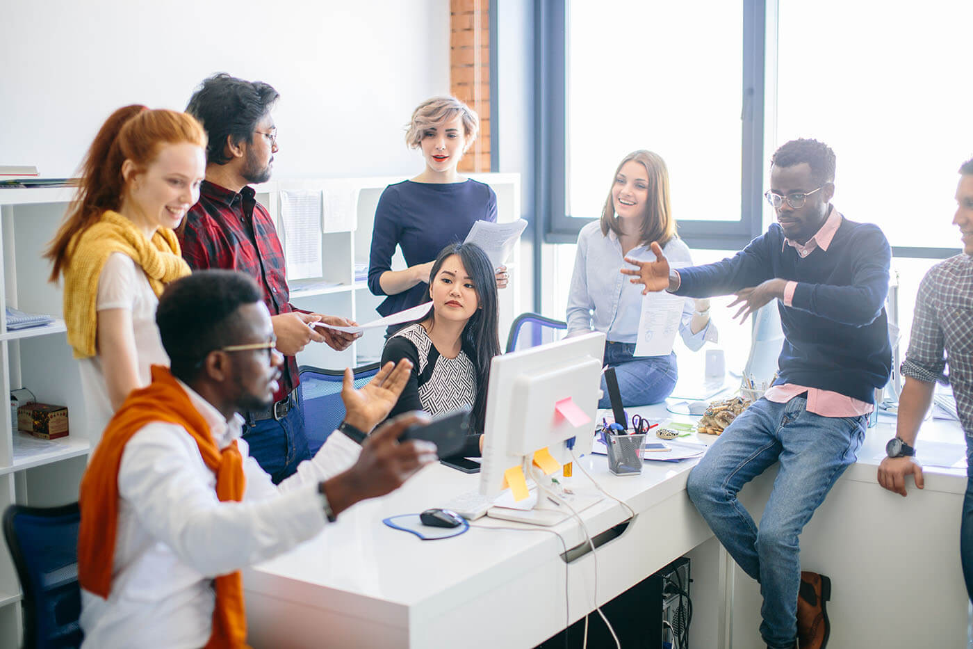 A diverse team of seven people engaged in a lively discussion around a computer in a bright, modern office setting.