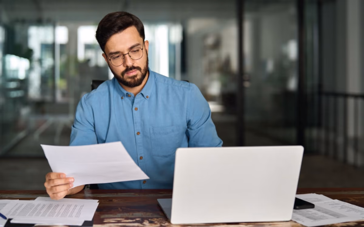 A man in a blue shirt and glasses sits at a desk, focused on a document in his hand while working on a laptop, conveying a studious atmosphere.