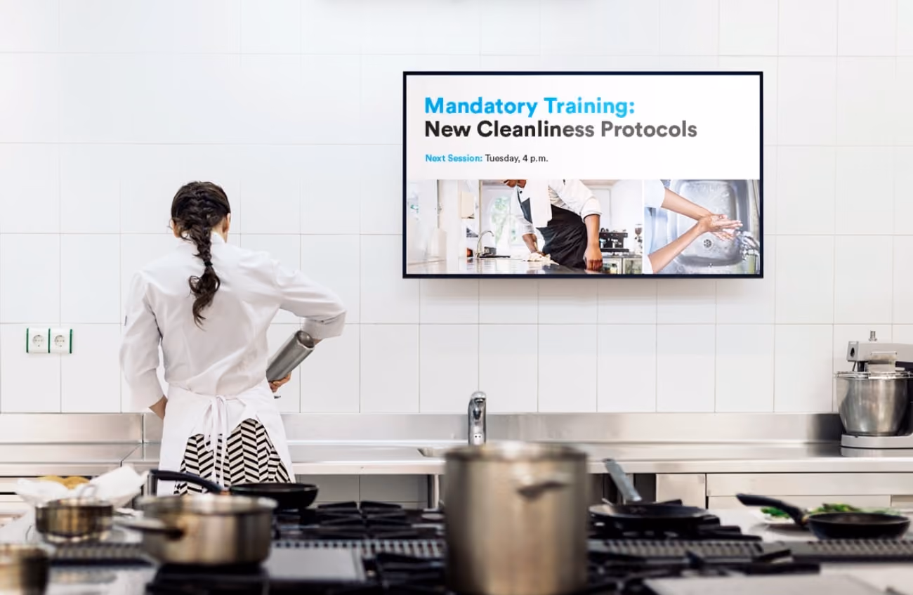 In a commercial kitchen, a chef in a white coat and patterned skirt pours from a metal container. A screen displays "Mandatory Training: New Cleanliness Protocols" with details.