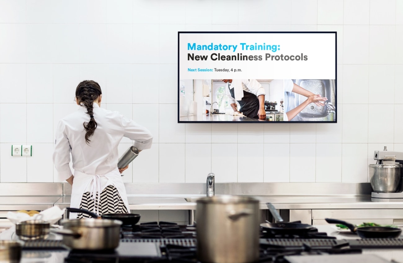 In a commercial kitchen, a chef in a white coat and patterned skirt pours from a metal container. A screen displays "Mandatory Training: New Cleanliness Protocols" with details.